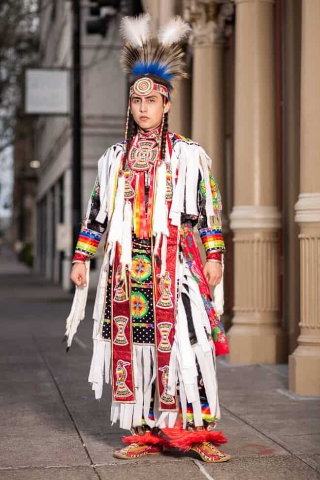 Young man in elaborate Native American powwow regalia with a feather headdress standing on a city sidewalk
