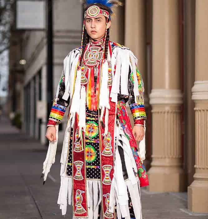 Young man in elaborate Native American powwow regalia with a feather headdress standing on a city sidewalk