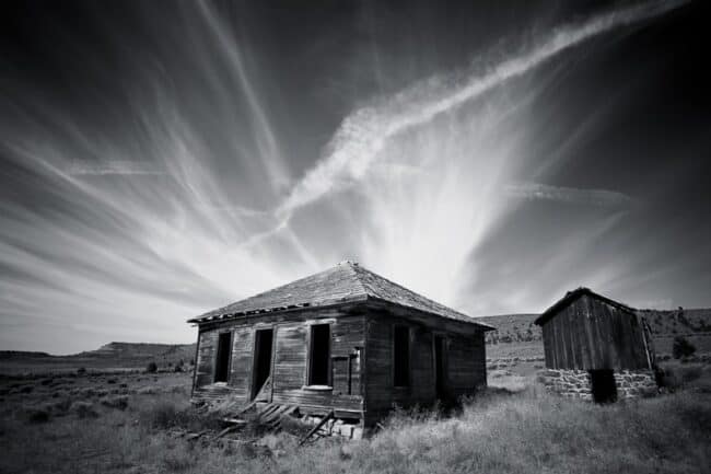 Black and white photo of an abandoned house and shed under a dramatic sky with a contrail