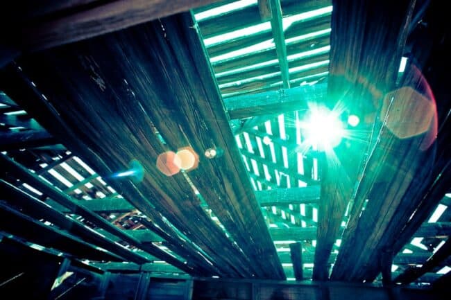 A low-angle view looking up through the rafters of a barn, with sunlight and lens flare streaming through gaps in the roof