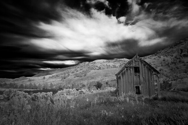 Black and white photo of an old wooden house beneath a dramatic stormy sky in the hills