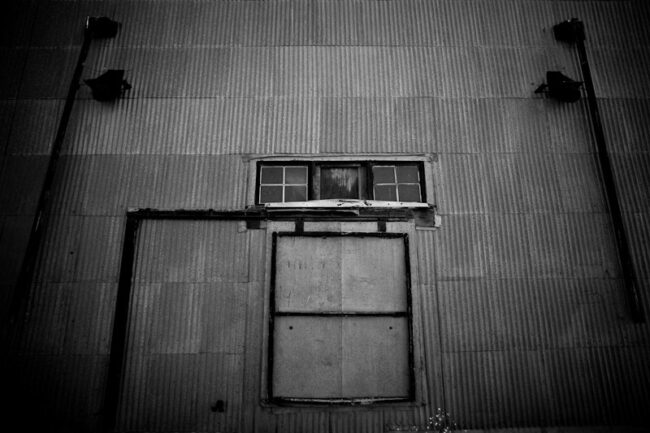 Black and white photo of a corrugated metal building facade with a boarded window and door