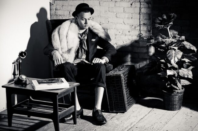 Black and white portrait of a man in a fur-trimmed coat and bowler hat seated in an armchair with a vintage telephone and suitcase