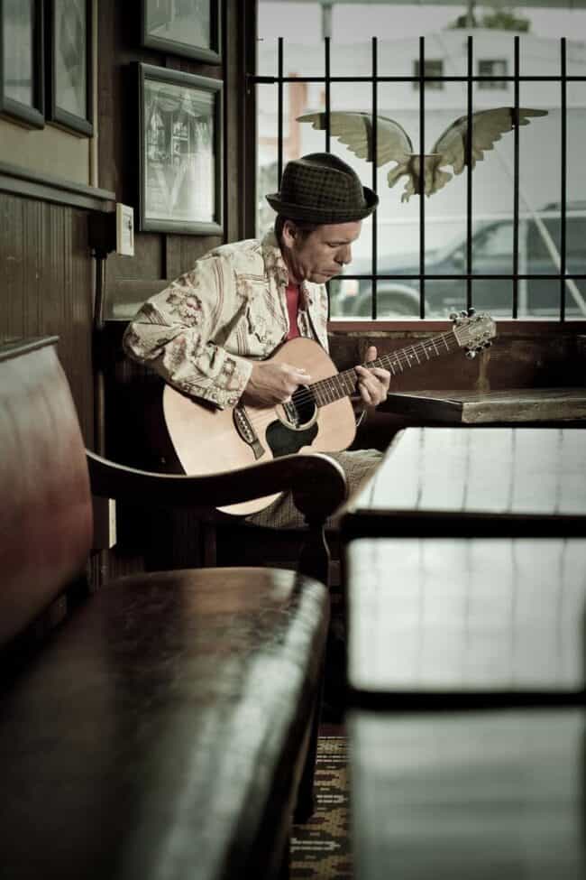 Man in a patterned jacket and fedora playing acoustic guitar in a cafe with angel wing decor