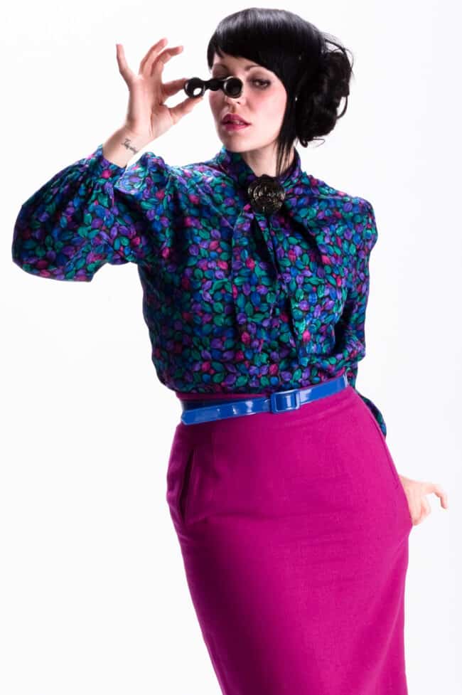 Woman in a colorful floral blouse and magenta skirt holding vintage binoculars in a studio portrait
