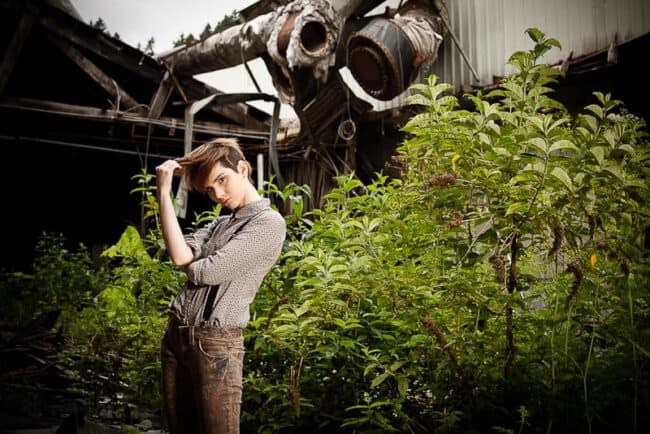 Woman with a pixie cut in a patterned shirt and overalls standing amid greenery near rusted industrial pipes