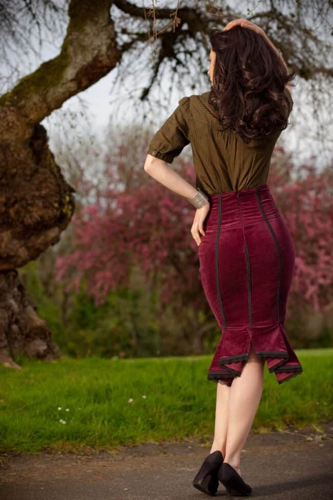 Woman seen from behind in an olive blouse and burgundy velvet pencil skirt in a garden with cherry blossoms