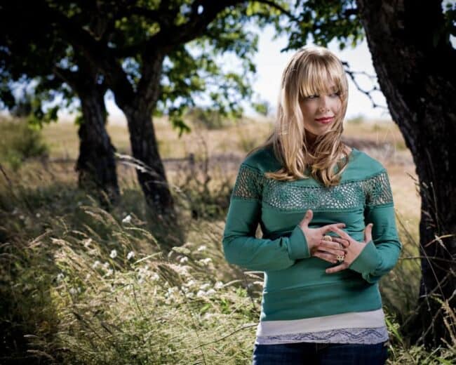 Blonde woman in a green top standing among trees with hands clasped