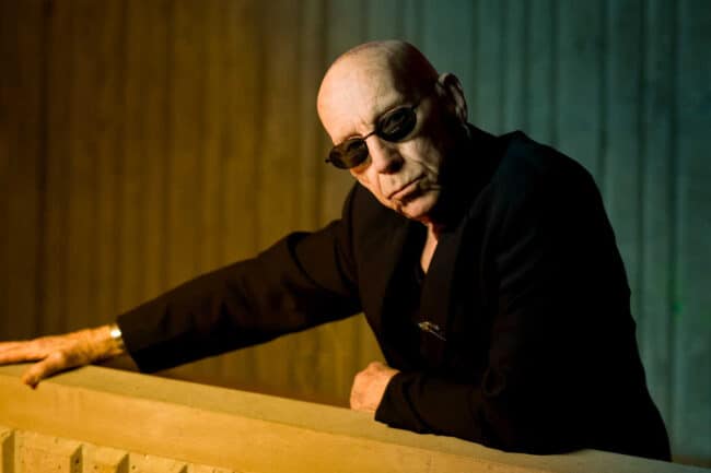Portrait of an older bald man in a black suit and sunglasses leaning on a railing