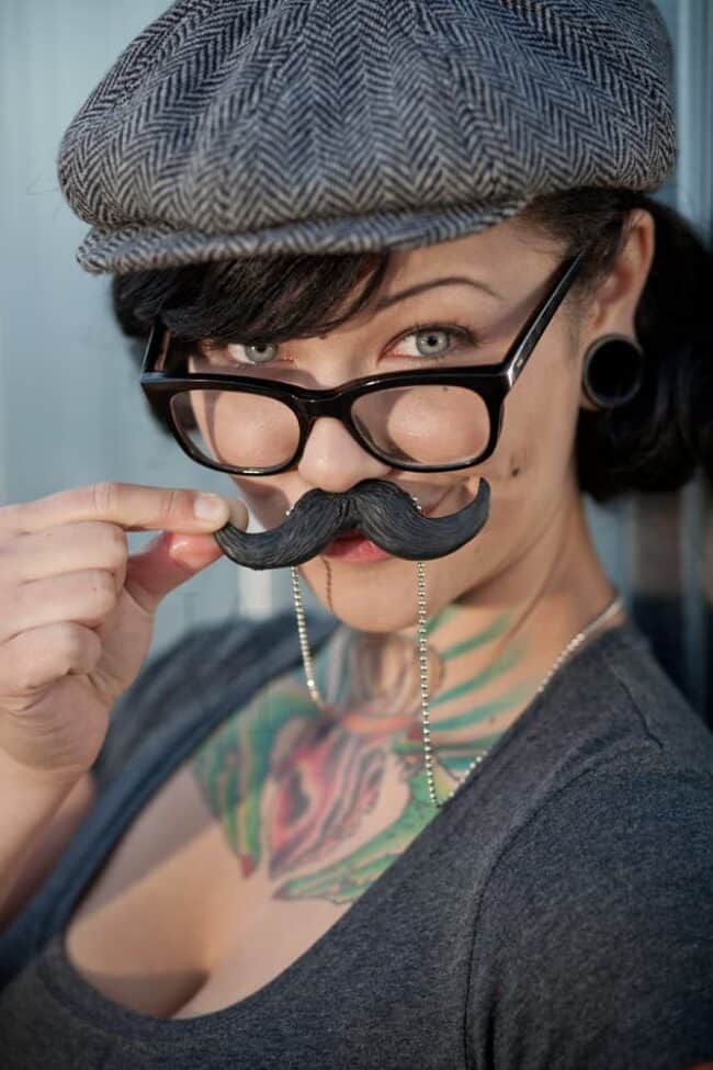 Woman in a newsboy cap and glasses holding a fake mustache to her face