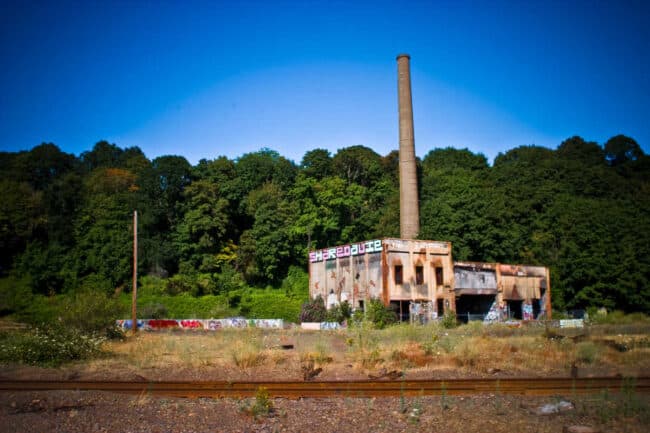 Abandoned graffiti-covered building with a tall smokestack beside railroad tracks