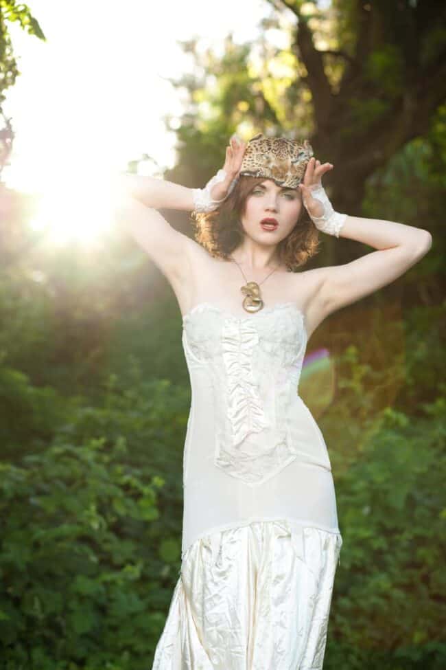 Woman in a white gown with a leopard print headpiece backlit by sunlight in a forest