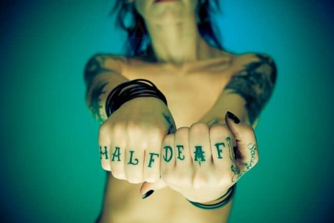 Close-up of a person's fists with 'HALF DEAF' tattooed across the knuckles, teal toned