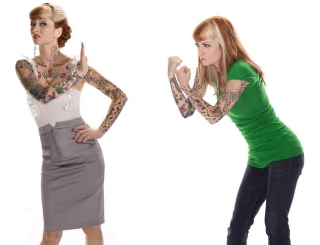 Two women with tattoo sleeves posing in a mock confrontation, one gesturing stop and one with fists raised