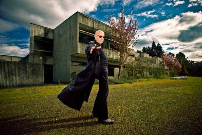 Man in a black coat and sunglasses striding across a lawn in front of an abandoned building under a dramatic sky