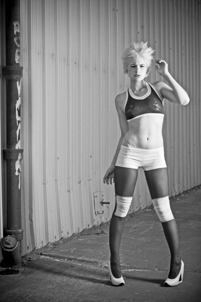 Black and white fashion portrait of a woman in a sporty crop top and shorts with sheer stockings against a corrugated metal wall