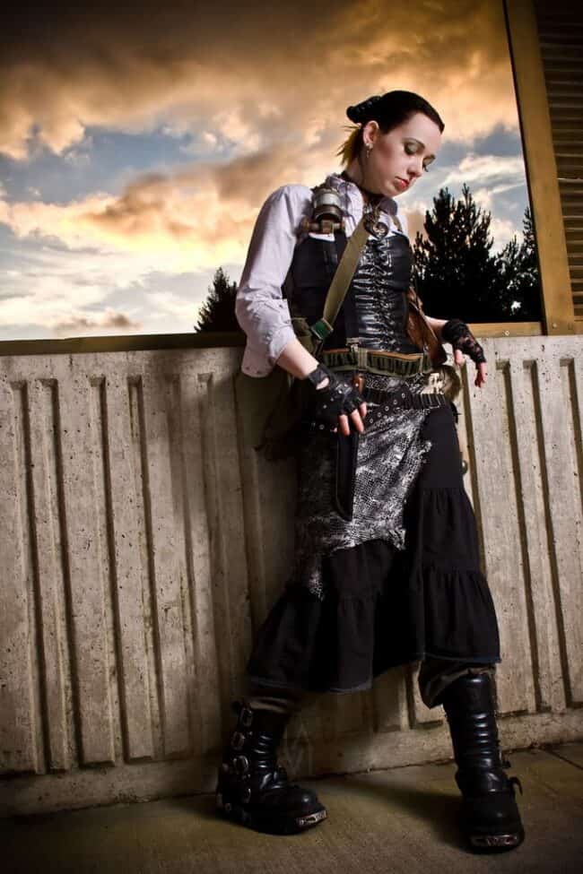 Woman in steampunk costume with goggles and vest leaning on a railing outdoors