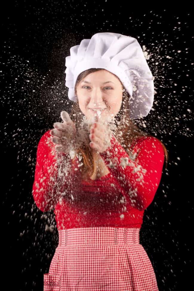 Woman in a chef hat and gingham apron with flour flying around her
