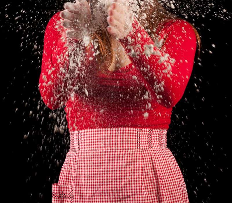 Woman in a chef hat and gingham apron with flour flying around her