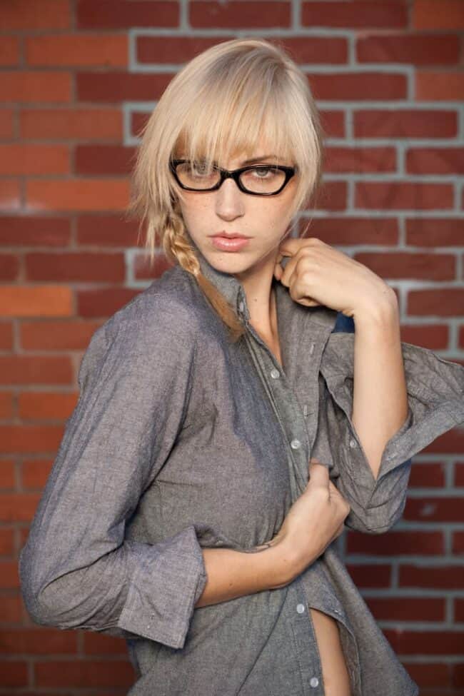 Fashion portrait of a blonde woman in glasses wearing an open gray shirt against a brick wall