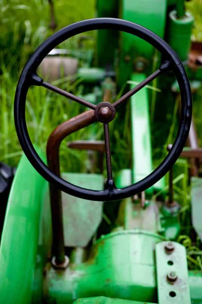 Close-up of a rusted steering wheel on old green farm machinery surrounded by grass