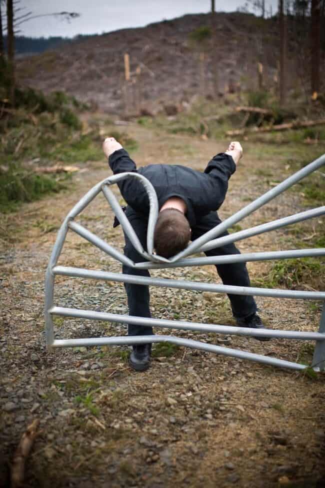 Man doing a handstand on a metal farm gate in a clear-cut forest landscape