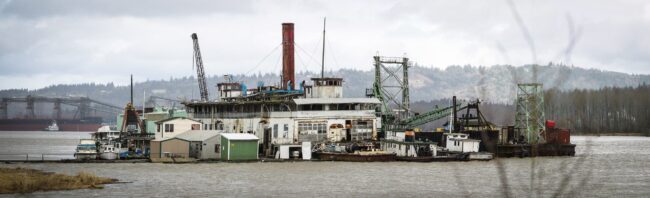 A weathered riverboat docked on the Columbia River with a bridge in the background