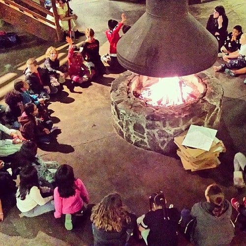 Group of children sitting in a circle around a lit outdoor fire pit at night