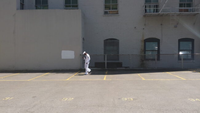 A person in a white uniform standing in an empty parking lot beside a brick building