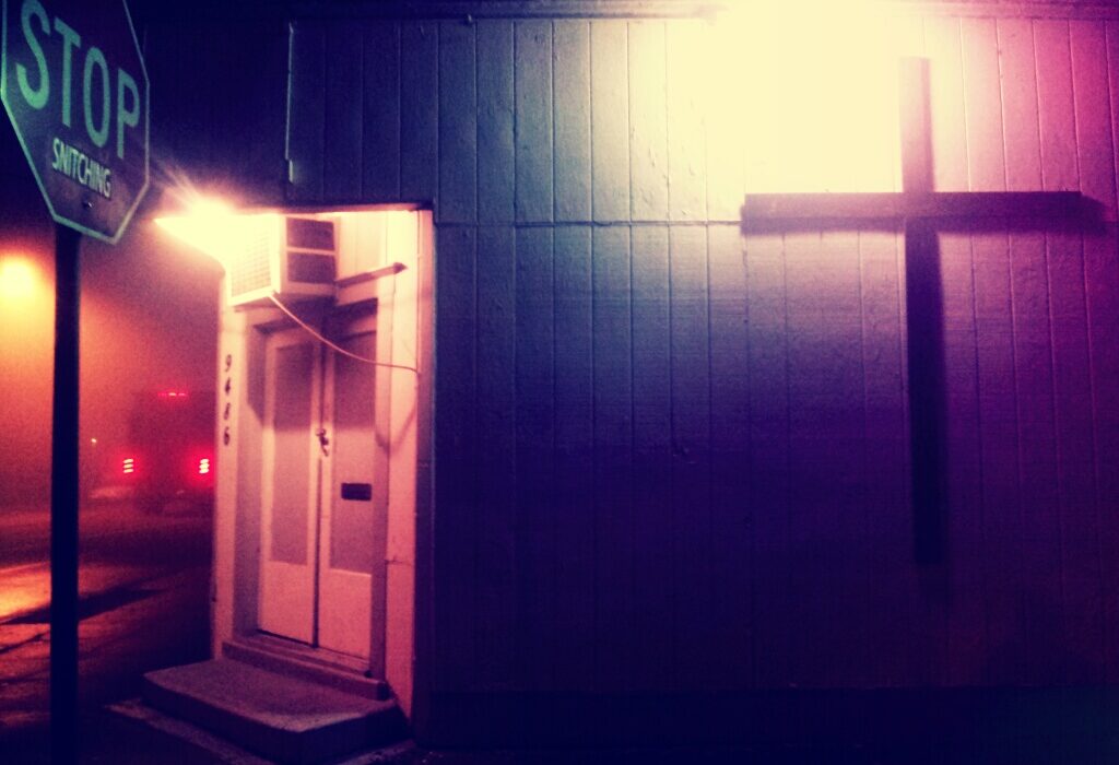 A lit cross on a building at night next to an altered stop sign