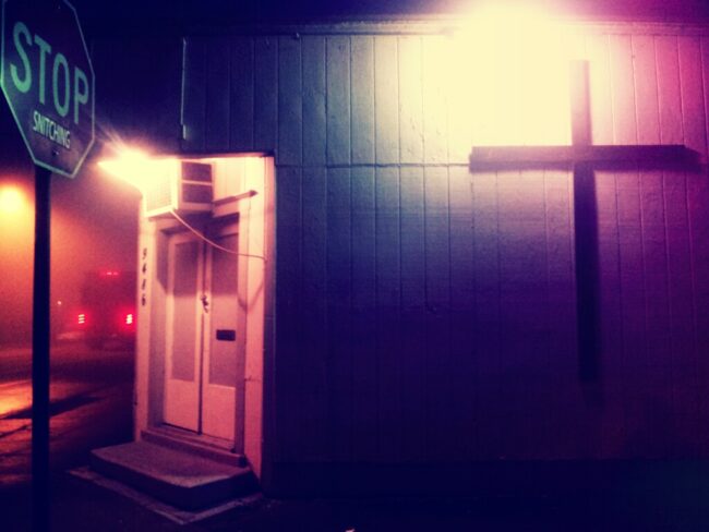 A lit cross on a building at night next to an altered stop sign