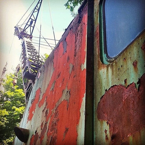 Split image of a rusted ship hull with peeling paint next to a rigging crane and trees