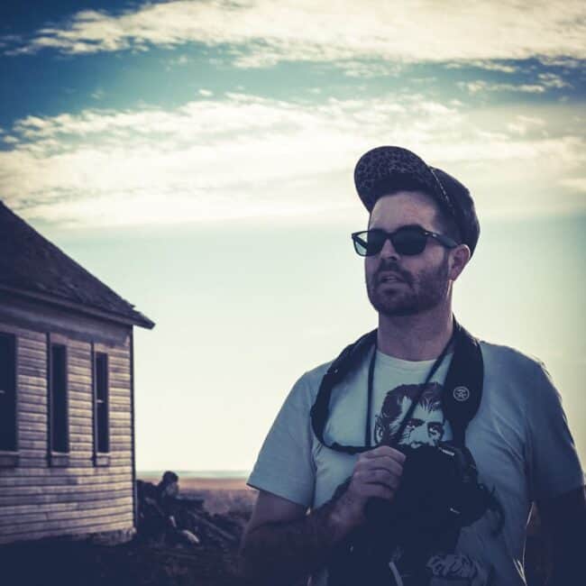 Photographer wearing a cap and sunglasses standing near an old prairie schoolhouse