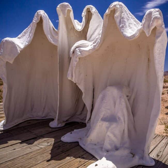 Three ghost-like white draped plaster figures on a wooden deck in the desert