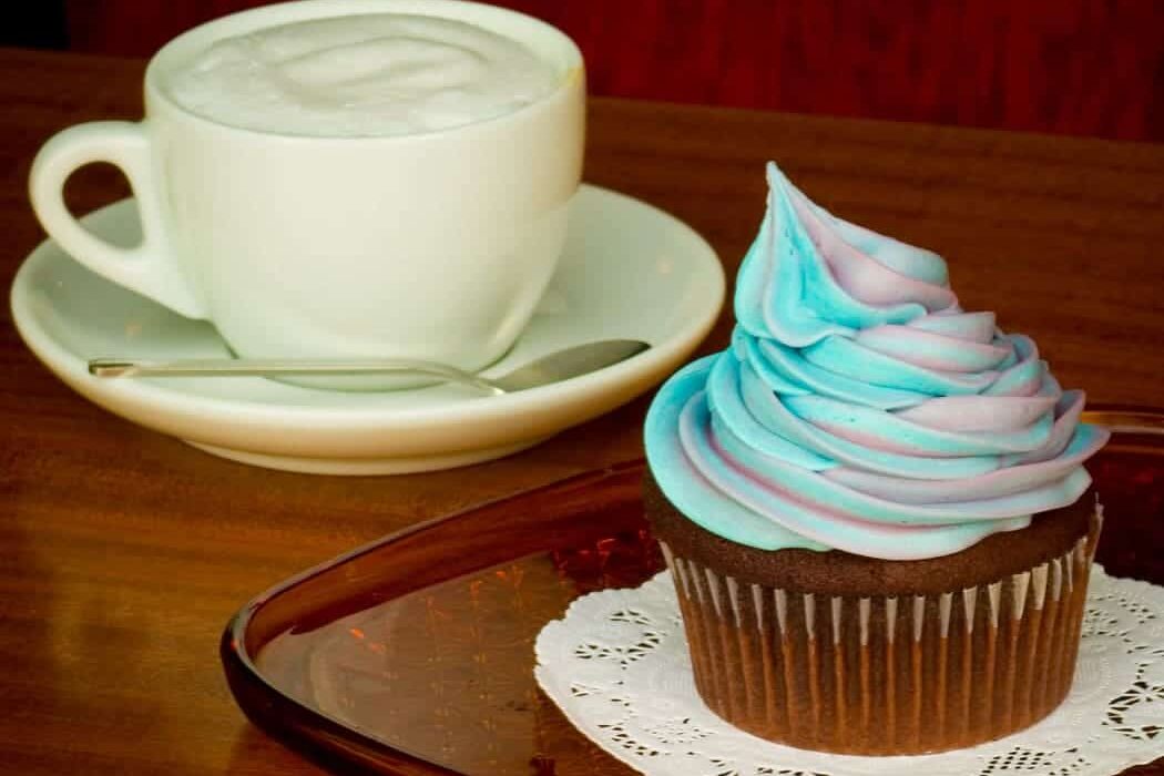 A frosted cupcake with swirled blue and purple icing beside a cappuccino on a wooden table