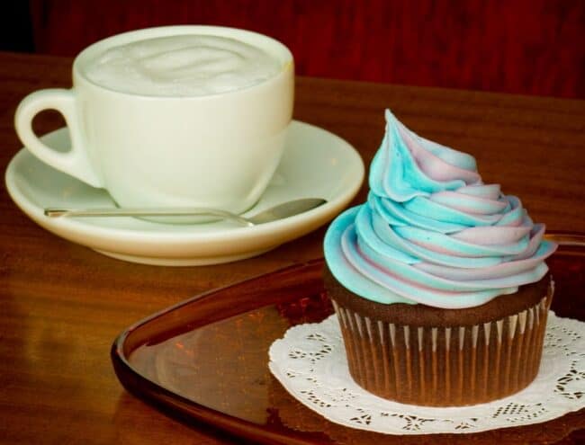 A frosted cupcake with swirled blue and purple icing beside a cappuccino on a wooden table