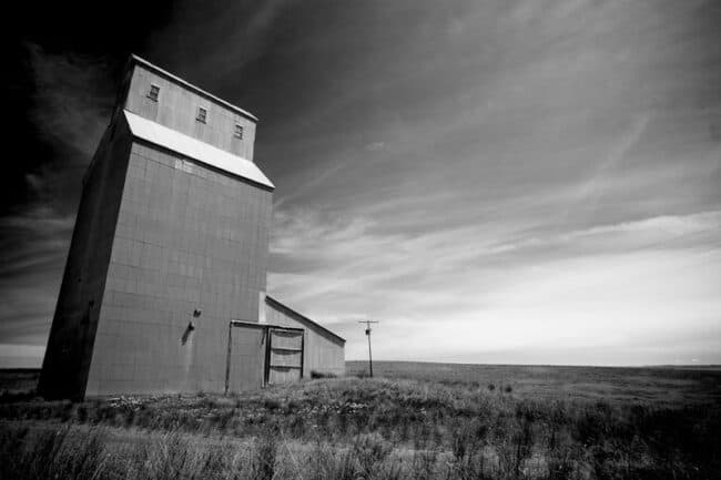 Black and white photograph of an old grain elevator standing in an open prairie landscape under a dramatic sky