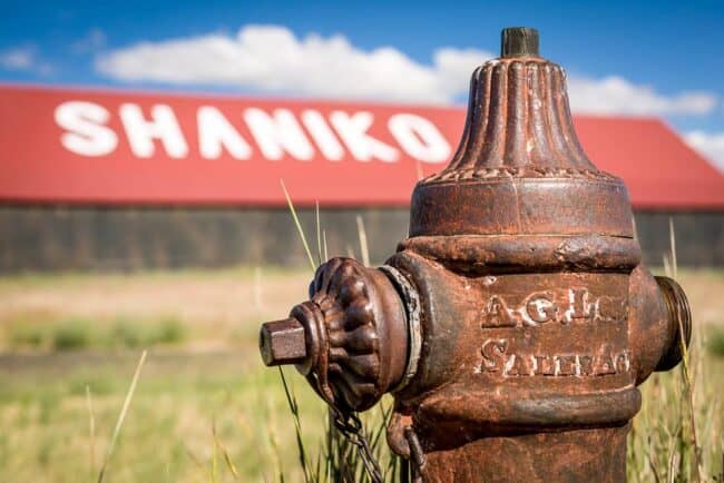 A close-up of a rusted antique fire hydrant with a red-roofed building reading "Shaniko" blurred in the background