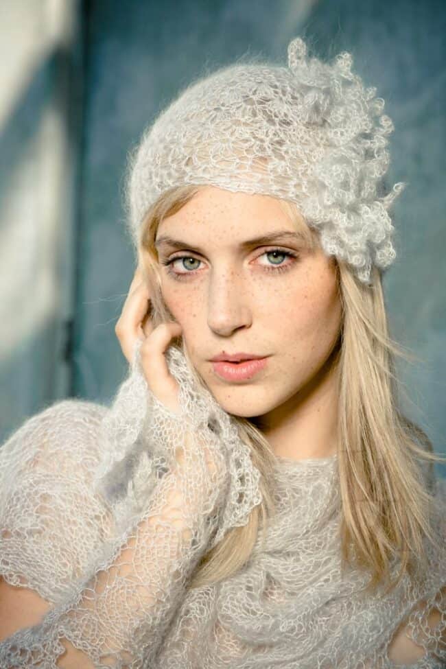 A woman with blonde hair and freckles wearing a white knit lace headpiece and wrap, hand resting near her face