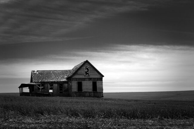 A black-and-white photo of an abandoned two-story farmhouse standing alone in an open field under a dramatic cloudy sky