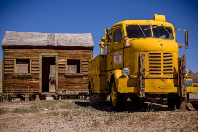 A vintage yellow water-well service truck parked next to a weathered wooden building