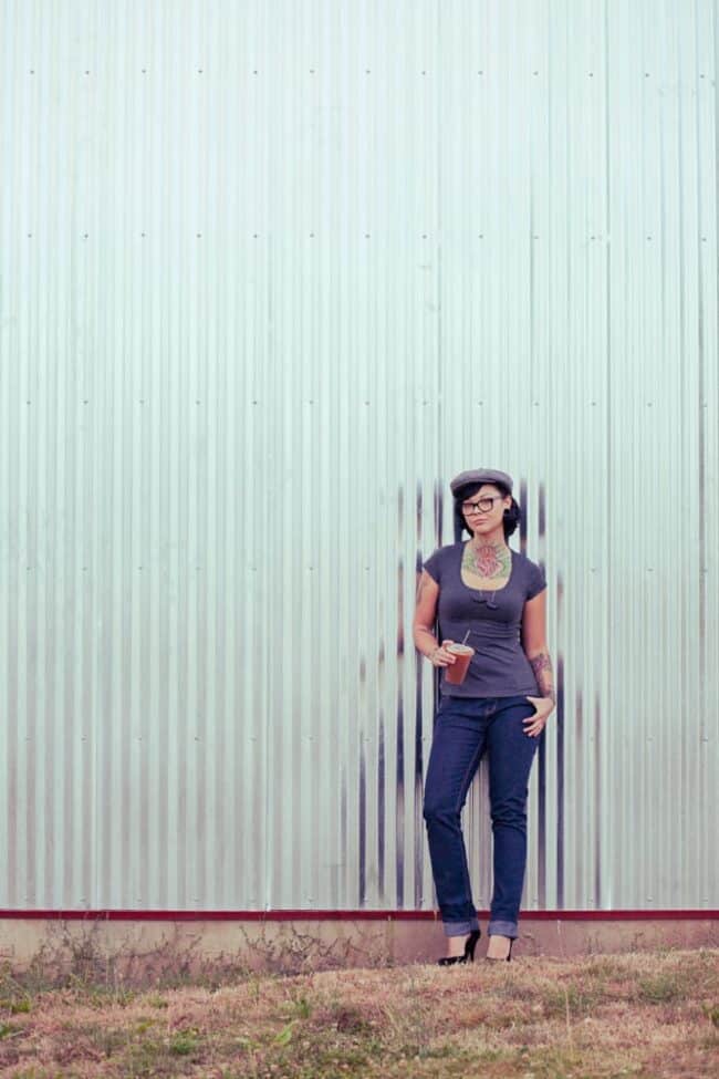 A woman wearing a newsboy cap and glasses, with visible tattoos, standing against a corrugated metal wall holding an iced drink