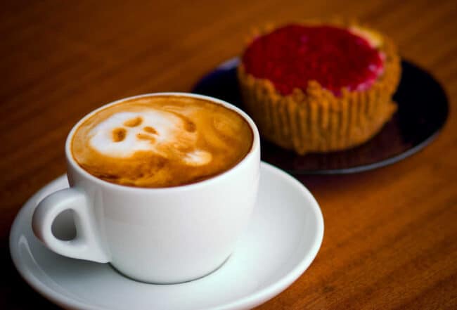 A cappuccino with a skull-shaped latte art design next to a raspberry-topped pastry on a wooden table
