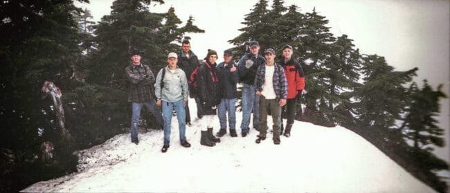 A vintage color photo of a group of hikers standing together on a snowy mountain ridge, surrounded by evergreen trees