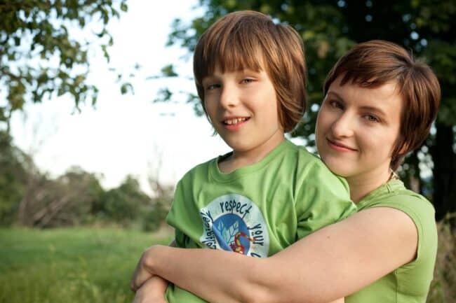 A woman with short brown hair smiling and hugging a young boy from behind in a grassy park, both wearing green shirts