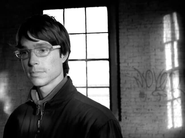 A black-and-white portrait of a young man wearing glasses standing near a large window, graffiti visible on the brick wall behind him