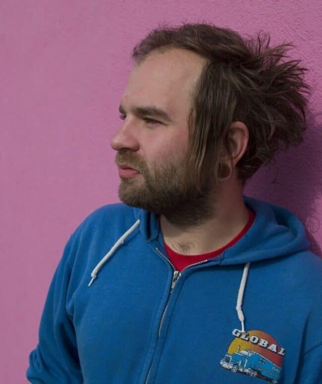 A profile portrait of a man with a wind-blown half-shaved hairstyle and beard, wearing a blue zip-up hoodie, against a pink wall