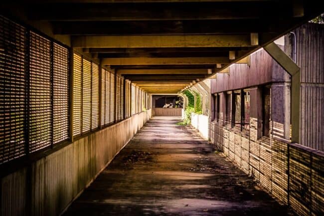 A long covered walkway with symmetrical perspective, weathered wood beams and slatted walls leading toward an abandoned building