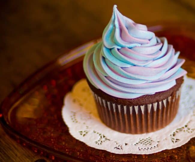 A chocolate cupcake topped with swirled pastel purple, pink, and blue frosting, on an ornate plate