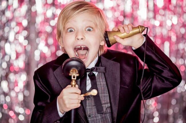 A young boy in a suit shouting excitedly into a vintage-style microphone, against a sparkly pink and silver background